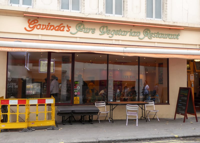 Beige restaurant facade with orange awning, large windows. Tables and chairs outside.