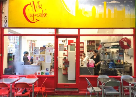 Red bakery facade with glass door and windows. Chairs and tables outside.