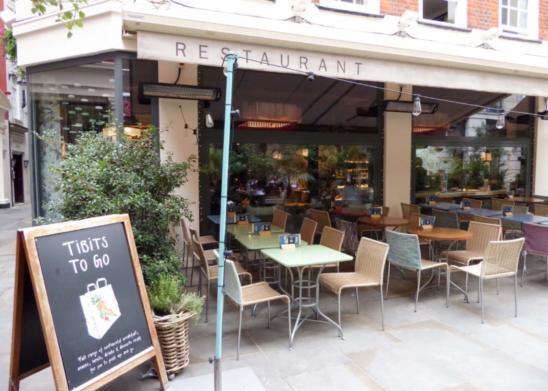 Beige restaurant facade with large windows. Table, chairs and sandwich board outside.