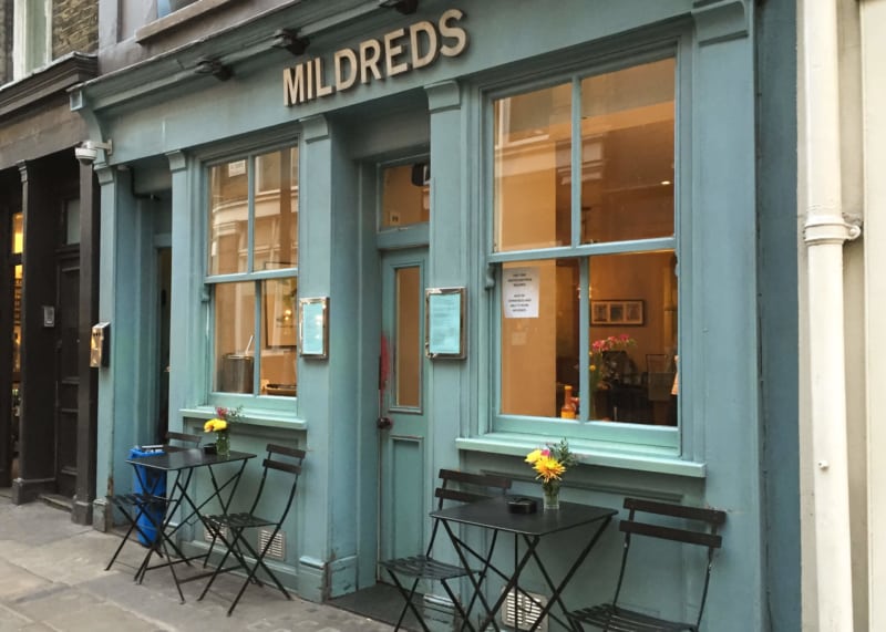 Turquoise restaurant facade with windows and door. Tables and chairs outside.