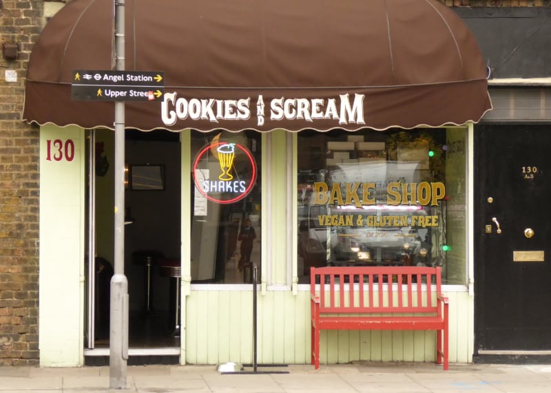 Green bakery facade with brown awning, large windows and open door. Red bench in front.